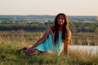 a young girl sitting on a grassy hill in front of a lake